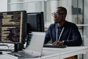 A man wearing glasses and a dark shirt works at a desk with two monitors and a laptop; one monitor displays cybersecurity code. He appears focused in a modern office setting with glass walls.